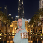 Woman in a light green hijab and dress holding a gold handbag in front of a city skyline at night.